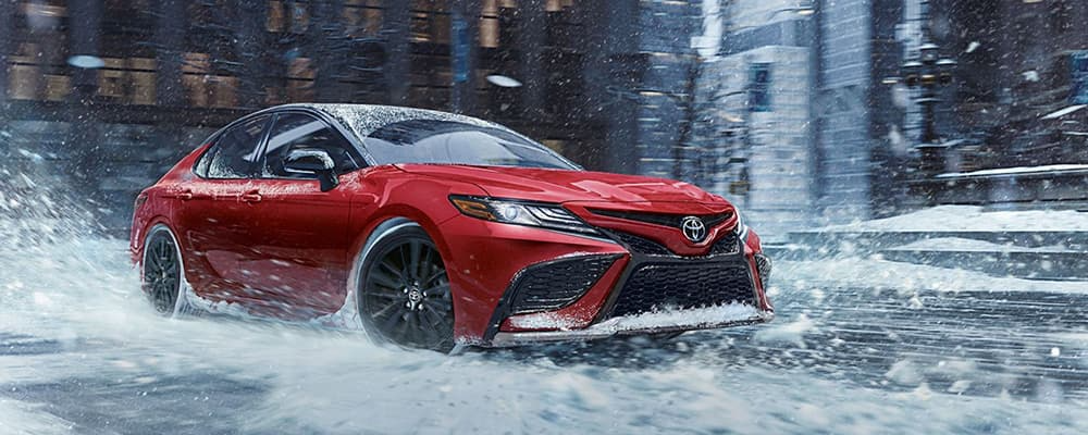 A red Toyota Camry drives on a snow covered, city street.