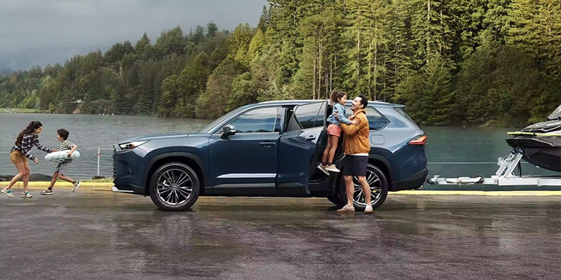 A man lifts his daughter out of a blue 2024 Toyota Grand Highlander parked near a lake.